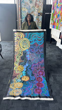 Load image into Gallery viewer, Rhoda behind her large colourful artwork displayed vertically in an indoor gallery painting features an abstract, patterned design with concentric circles, wavy lines, and bright colors including blue, yellow, orange, pink, purple, and black, with a white border at the bottom.

